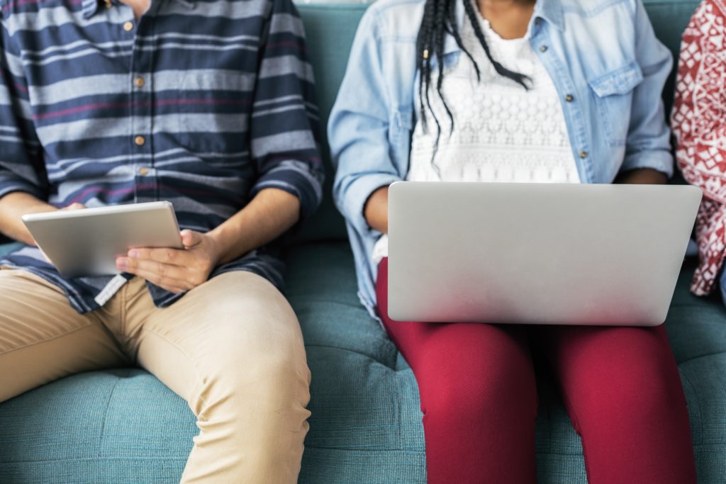 Two people sitting on a sofa, one person holding a tablet and the other a laptop. You can only see their torsos and legs, not their faces